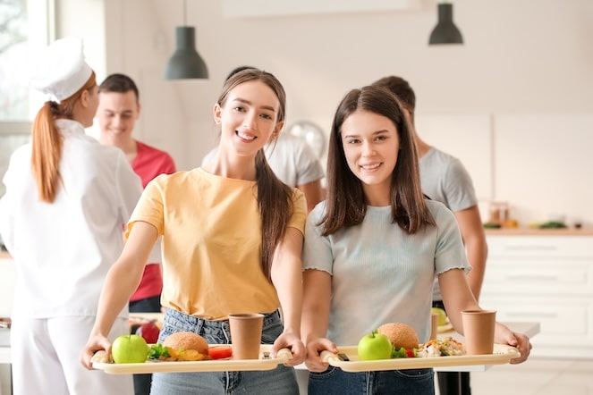 Two girls in the cafeteria holding very healthy lunches and smiling