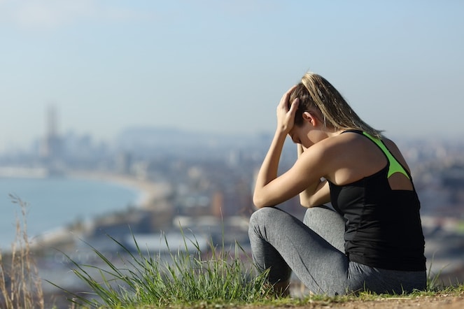 Depressed female athlete sitting on the side of a hill overlooking a city with her hands on her head. 