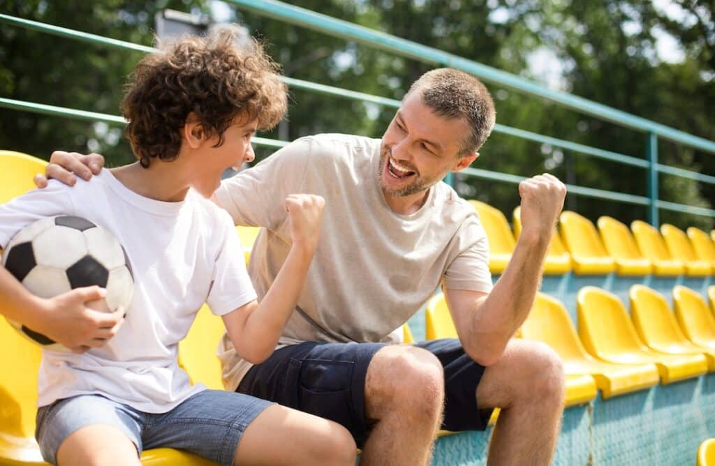 Dad sitting in the stands with his son, who is holding a soccer ball, very excited about something.
