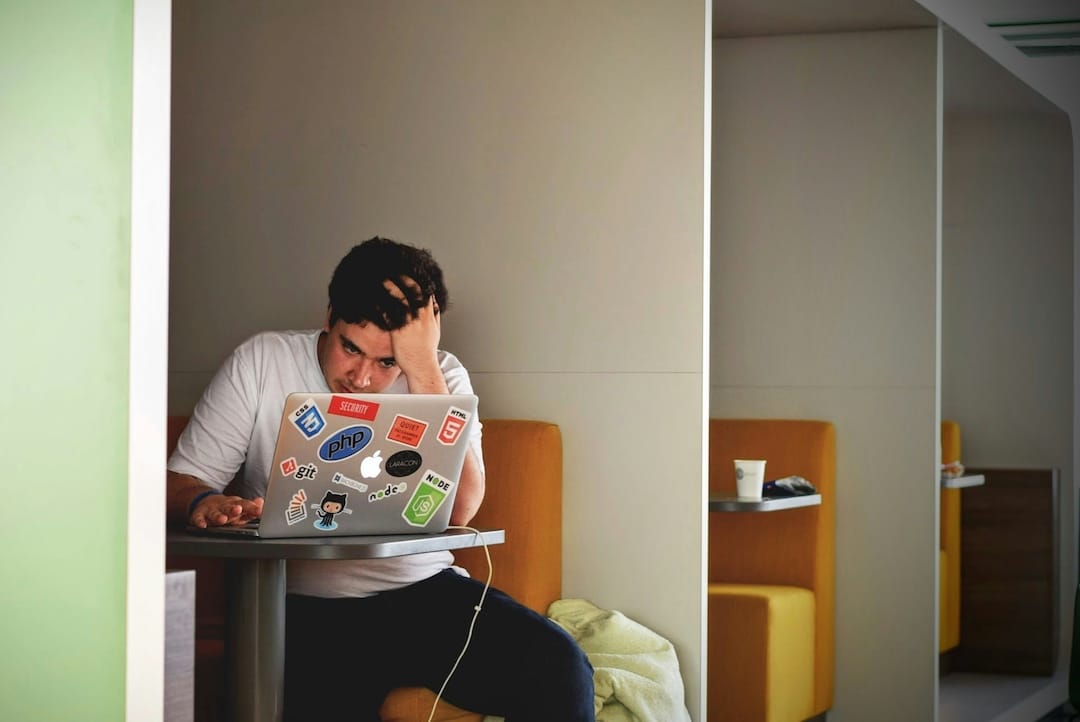 Confused young man with hand on head sitting an a booth with an open laptop.